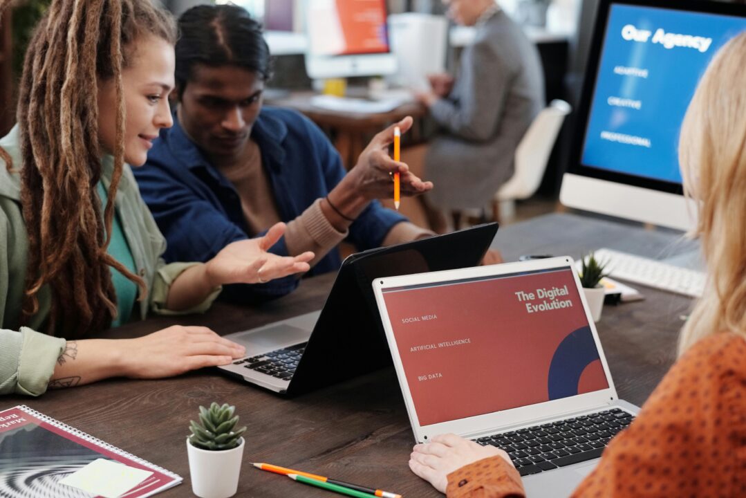 Group of young professionals collaborating at a modern office table, discussing ideas with laptops open, one screen displaying "The Digital Evolution" presentation.