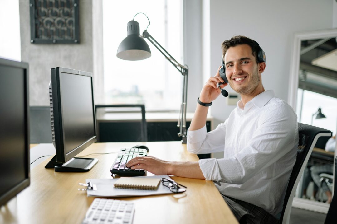 Smiling guy wearing a headset, sitting at a desk with dual monitors in a modern office environment.